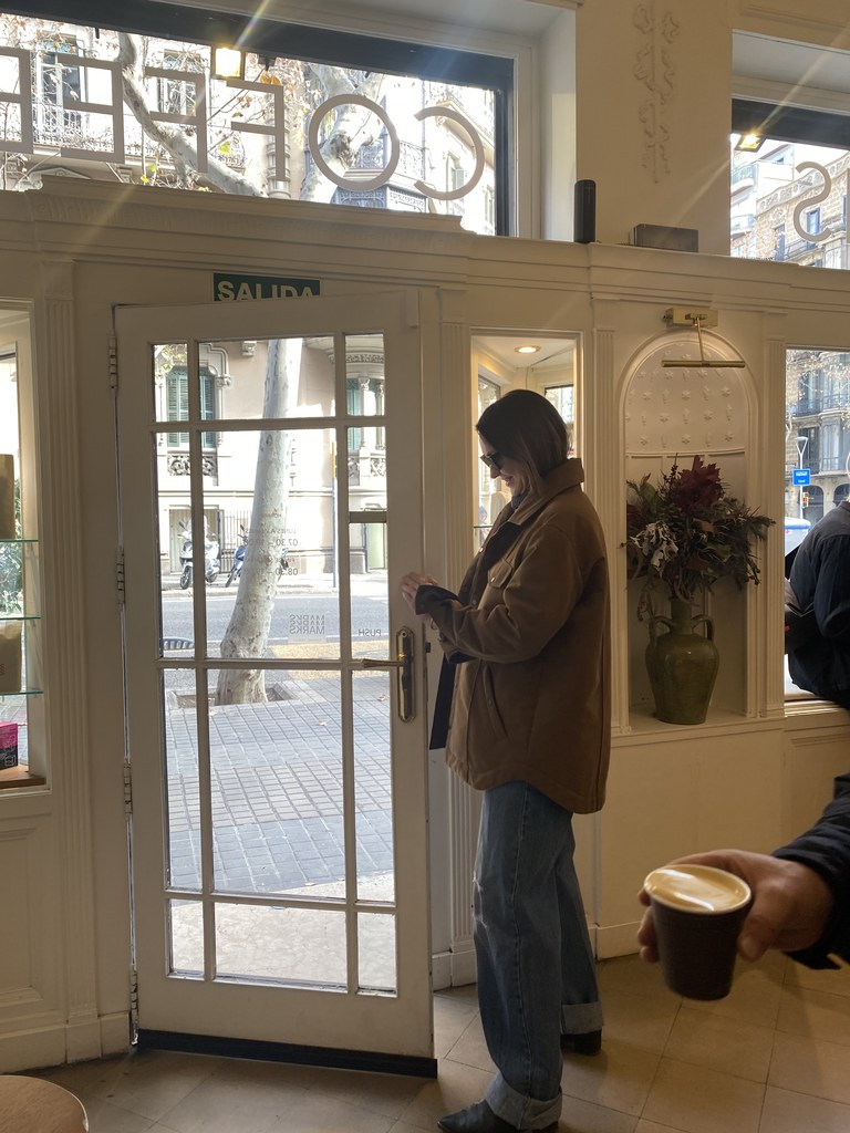 Девушка в коричневой куртке стоит у стеклянной двери в кафе, рука с кофе на переднем плане