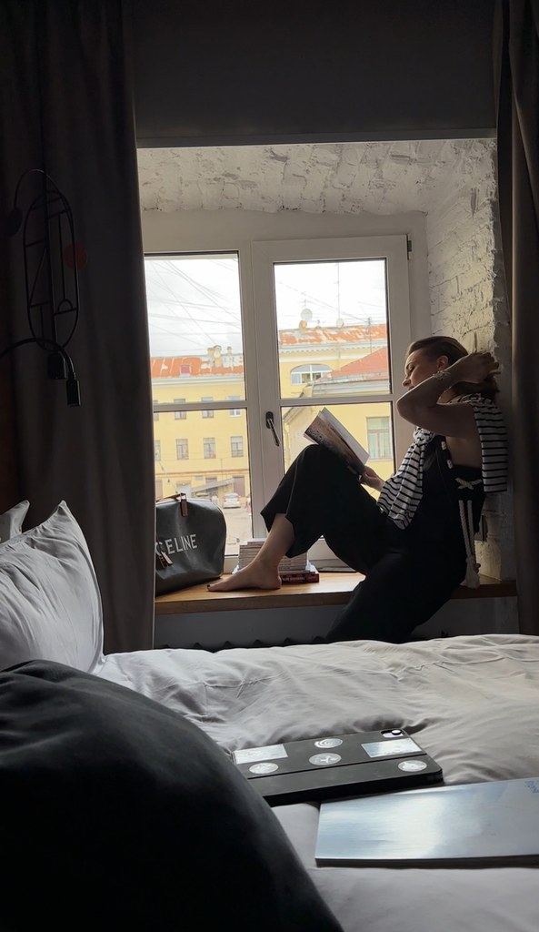 Девушка сидит на подоконнике у окна и читает книгу в тёмном номере